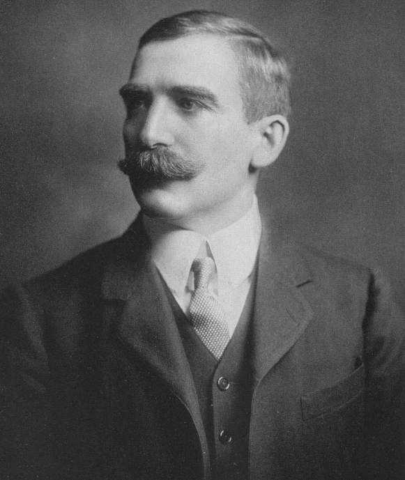 Sir henry Wellcome in a black and white image wearing a coat and tie with a mustache