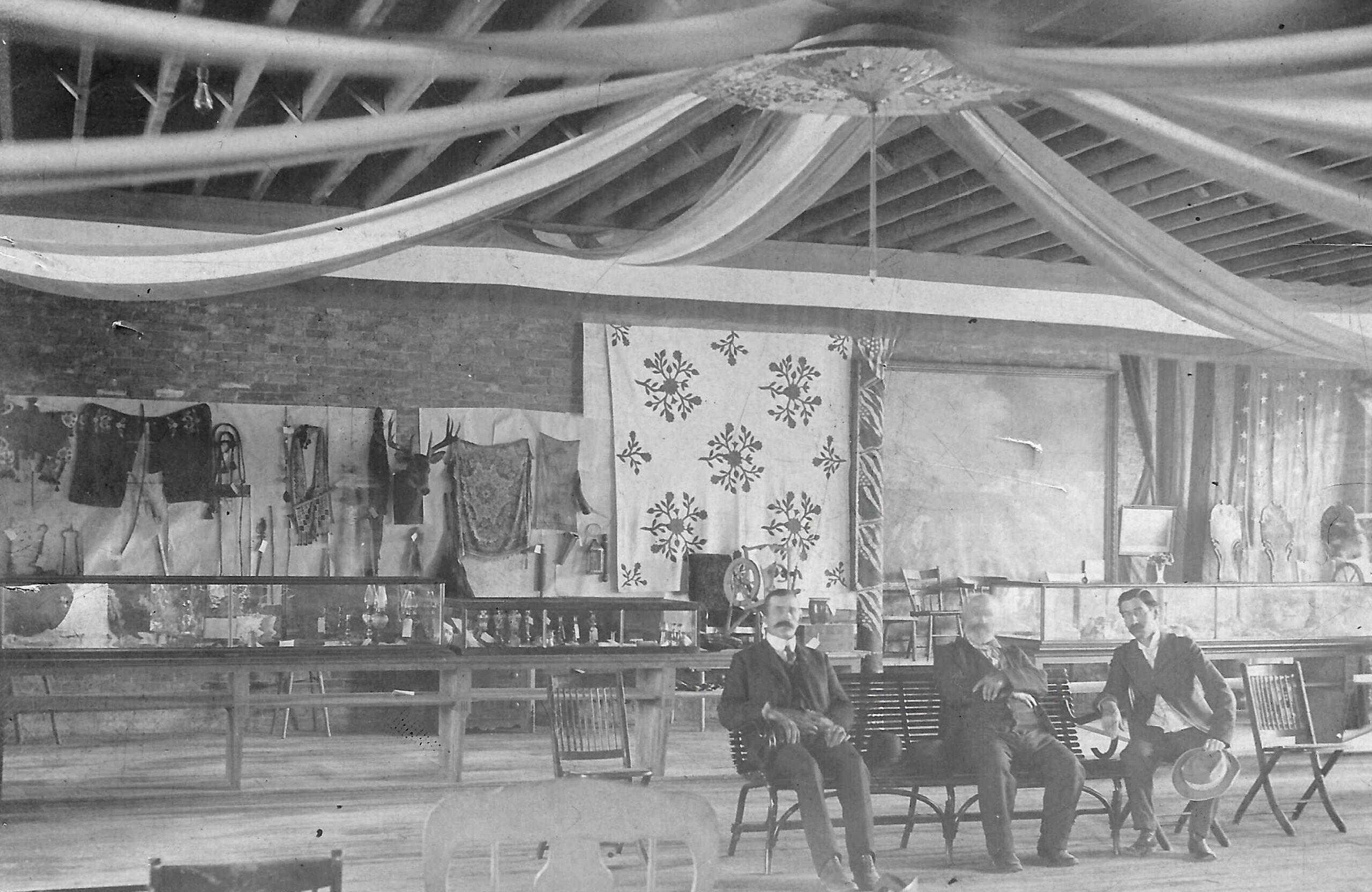 Black and white image of three guys in the Sibley Park Pavilion with items on display behind them